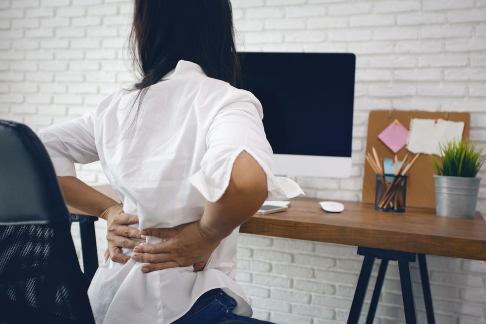 Young woman at desk holding her back in pain