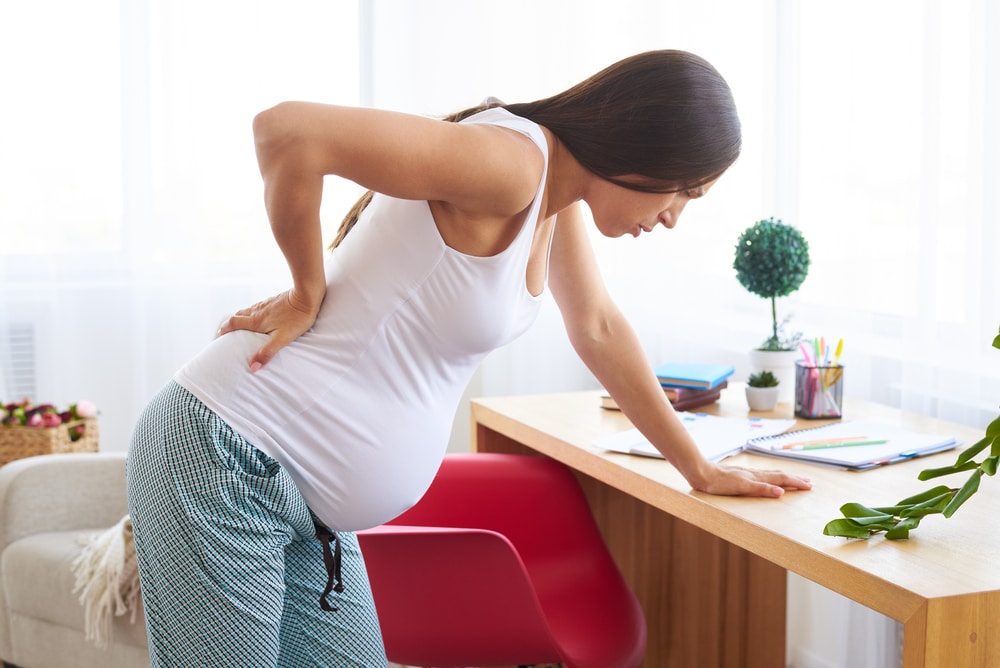Pregnant woman bent over desk with hand on back in pain